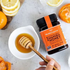 Wedderspoon Raw Manuka honey KFactor 16 jar with honey dipper and citrus fruits on a white surface