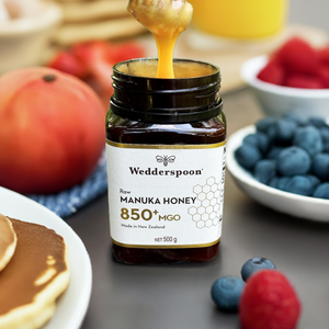 Jar of Wedderspoon Raw Manuka honey 850 MGO 500g being drizzled over pancakes in a breakfast setting with berries