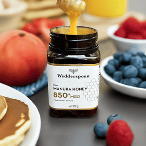 Jar of Wedderspoon Raw Manuka honey 850 MGO 500g being drizzled over pancakes in a breakfast setting with berries