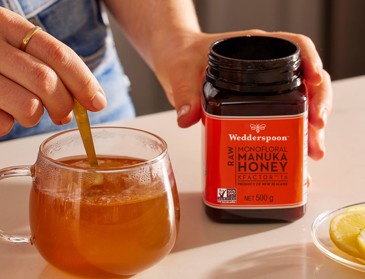 Hand stirring tea as jar sits beside mug with lemon slice Wedderspoon Manuka honey is an Essential, Lily Rabe .