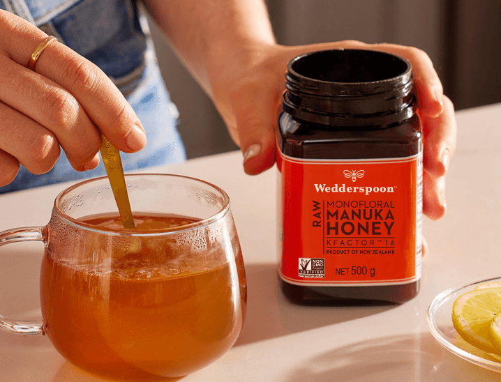Hand stirring tea as jar sits beside mug with lemon slice Wedderspoon Manuka honey is an Essential, Lily Rabe .
