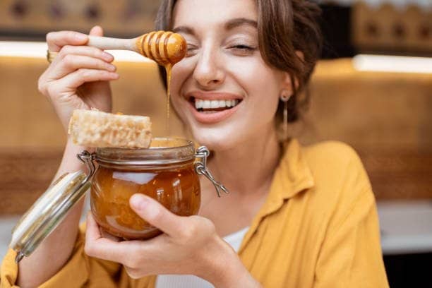 Smiling woman holding jar of Manuka honey and honeycomb tasting spoon while asking can diabetics eat manuka honey