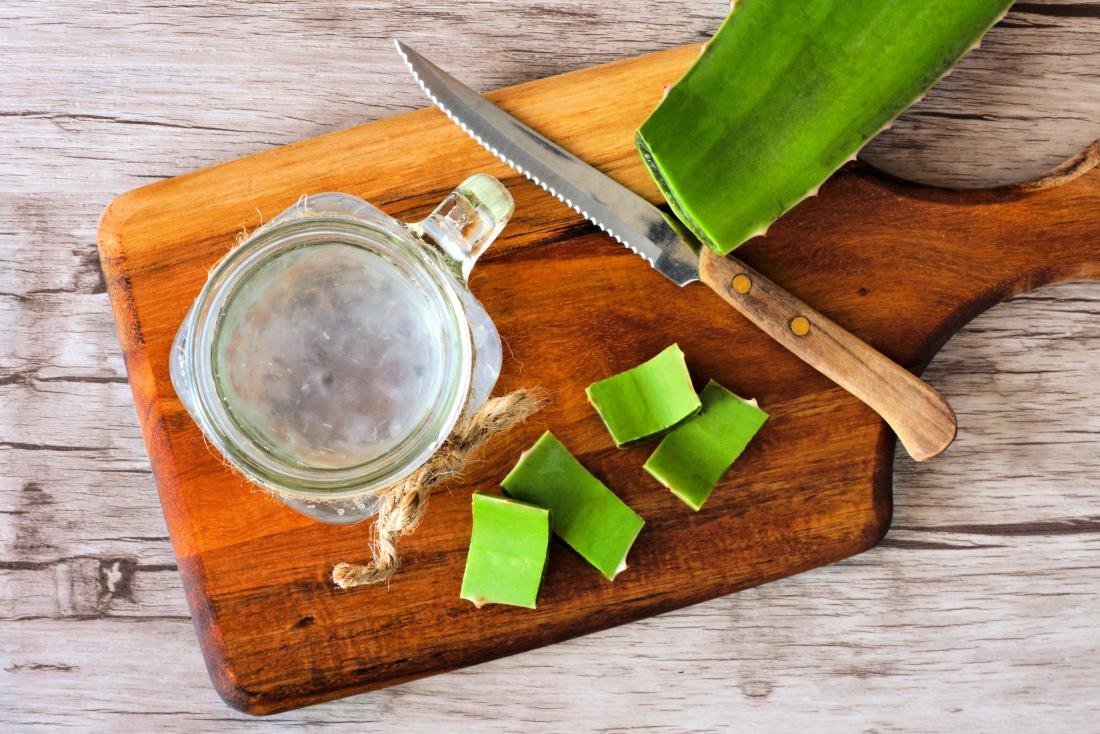 A jar of clear aloe drink with fresh aloe leaf pieces and knife on wooden board showing Aloe vera juice health benefits