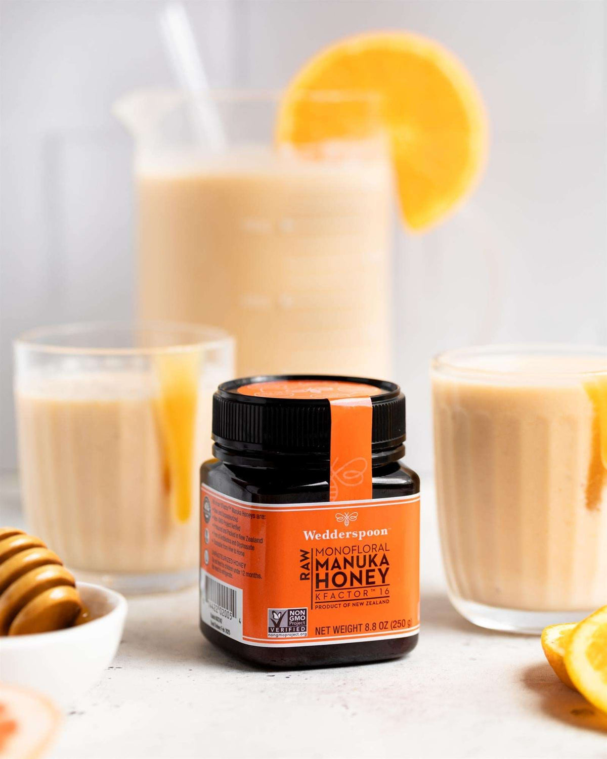 Wedderspoon Raw Manuka Honey 250g jar with creamy drinks and orange slices in background