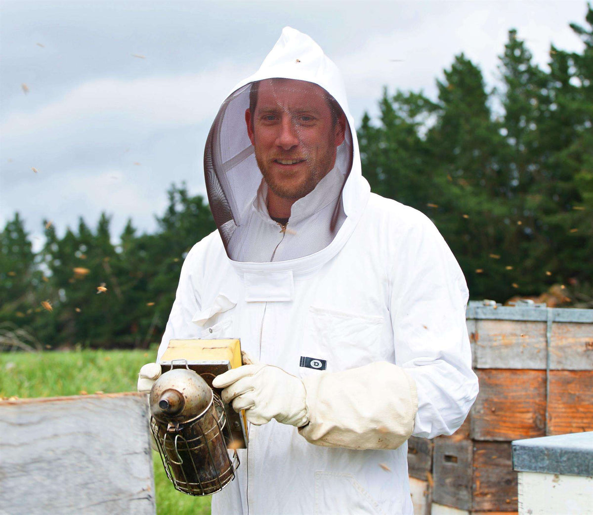 Beekeeper holding a smoker near beehives with Wedderspoon Raw Manuka Honey 250g in natural environment