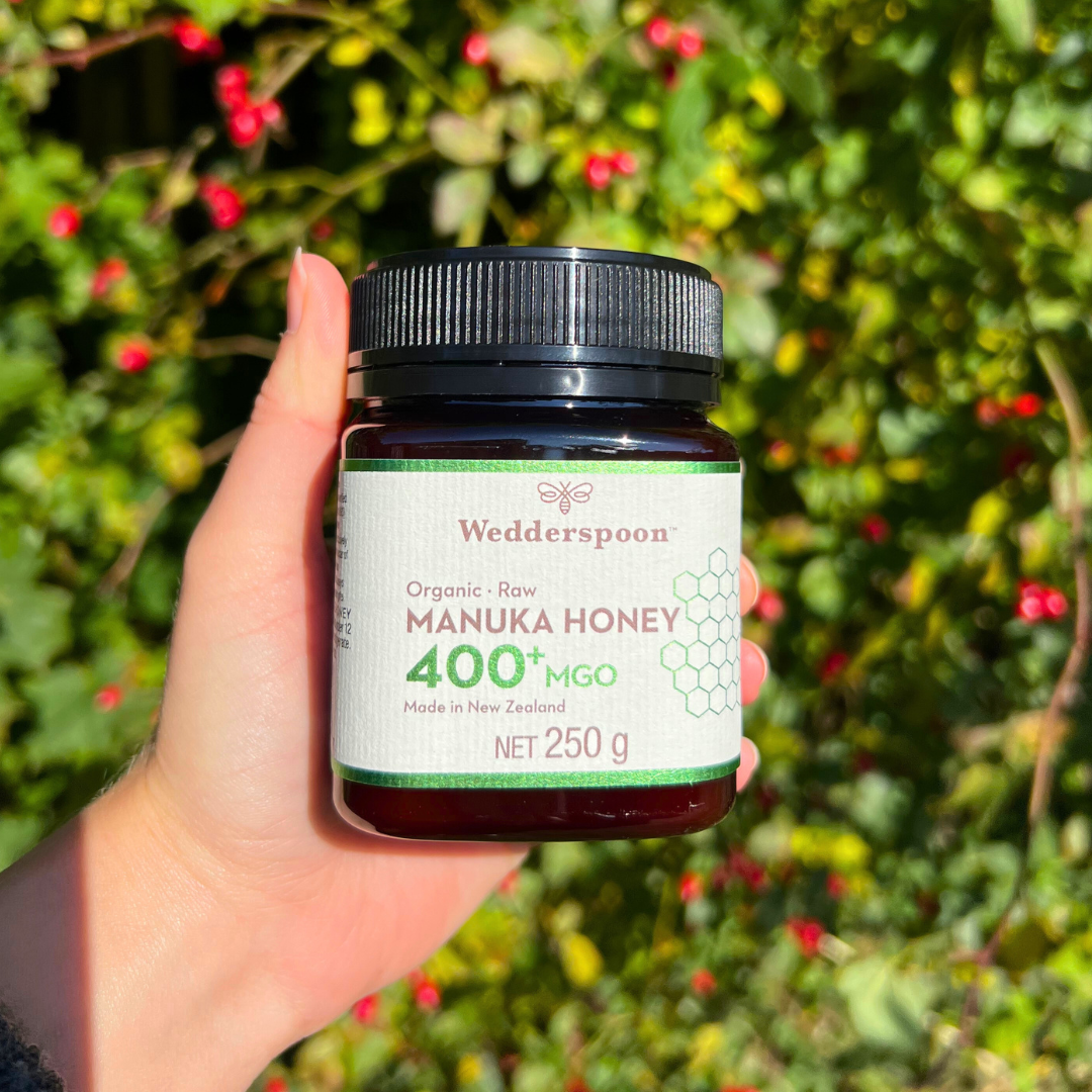 Wedderspoon Organic Raw Manuka Honey 400 MGO 250g jar held in hand with greenery background