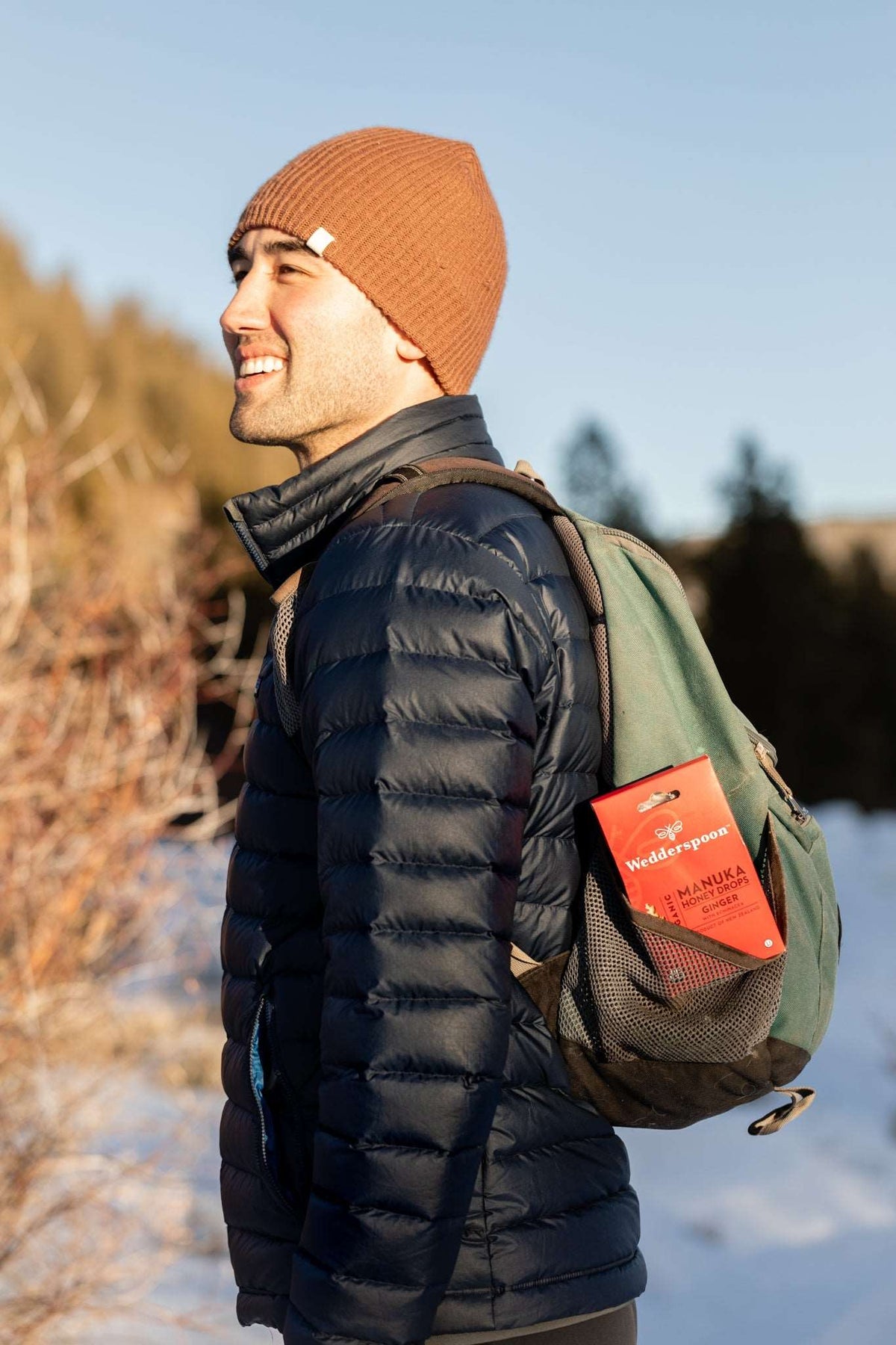 Man wearing brown beanie and black jacket carrying a green backpack with Wedderspoon manuka honey ginger drops throat lozenges in snow