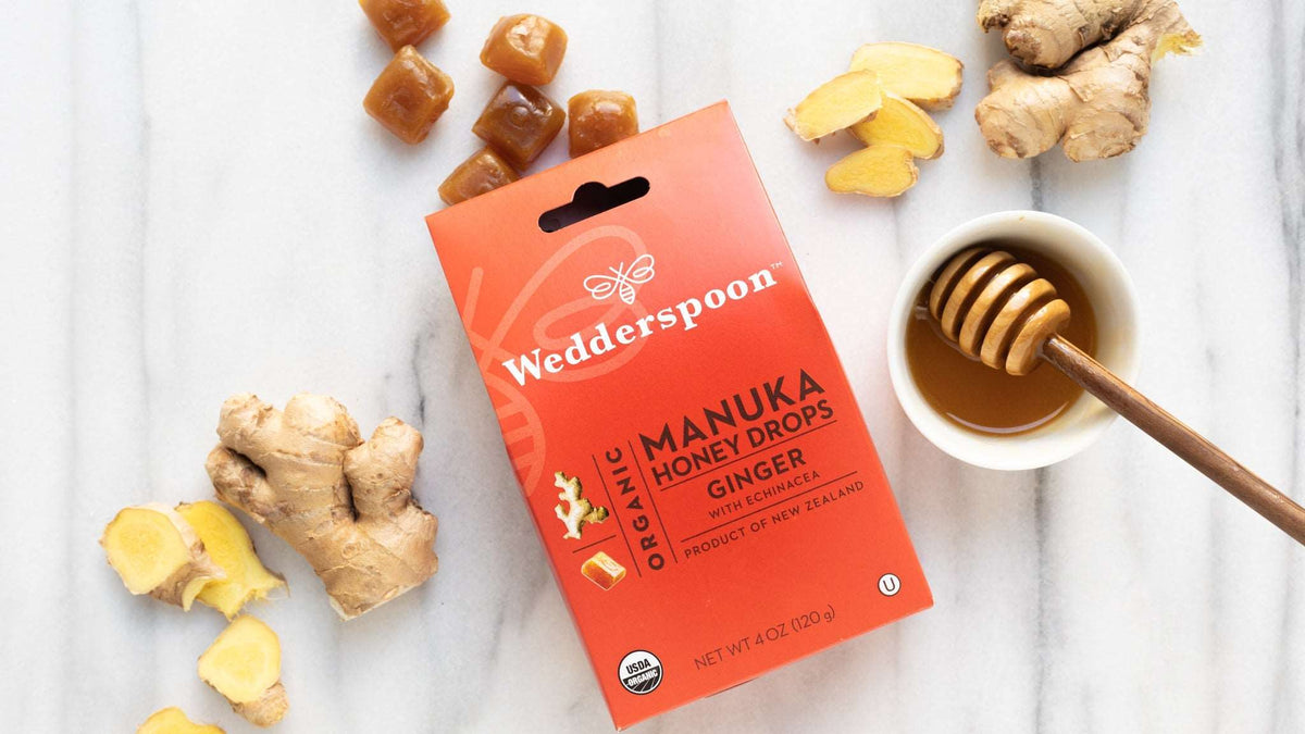Wedderspoon manuka honey ginger drops box with ginger root and honey dipper on white surface