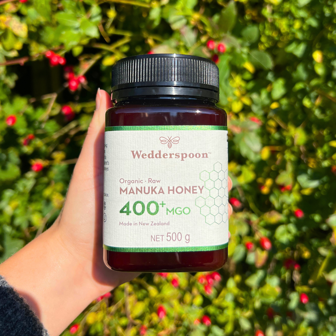 Wedderspoon Organic Raw Manuka Honey 400 MGO 500g jar held in hand with greenery background