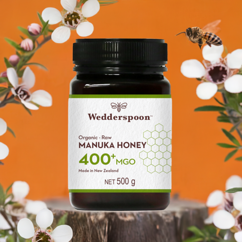 Wedderspoon Organic Raw Manuka Honey 400 MGO 500g jar with black lid on wooden surface with flowers and bee