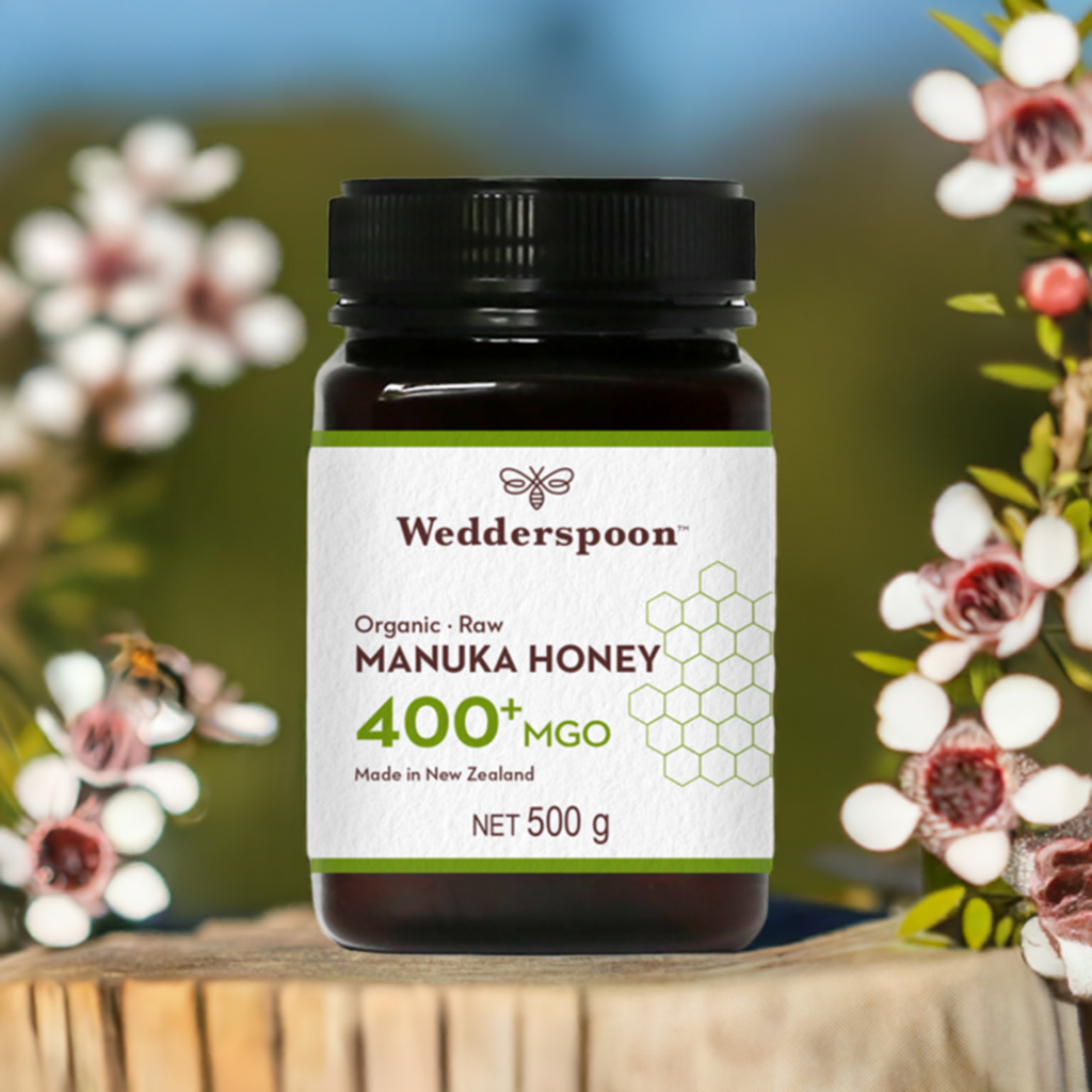 Wedderspoon Organic Raw Manuka Honey 400 MGO 500g jar on wooden surface with flowers background