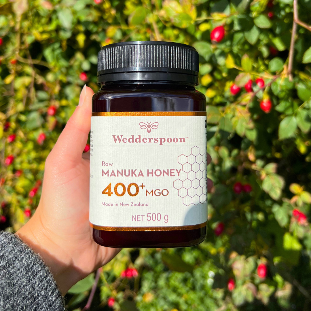 Wedderspoon Raw Manuka Honey 400 MGO 500g jar held outdoors with green foliage background