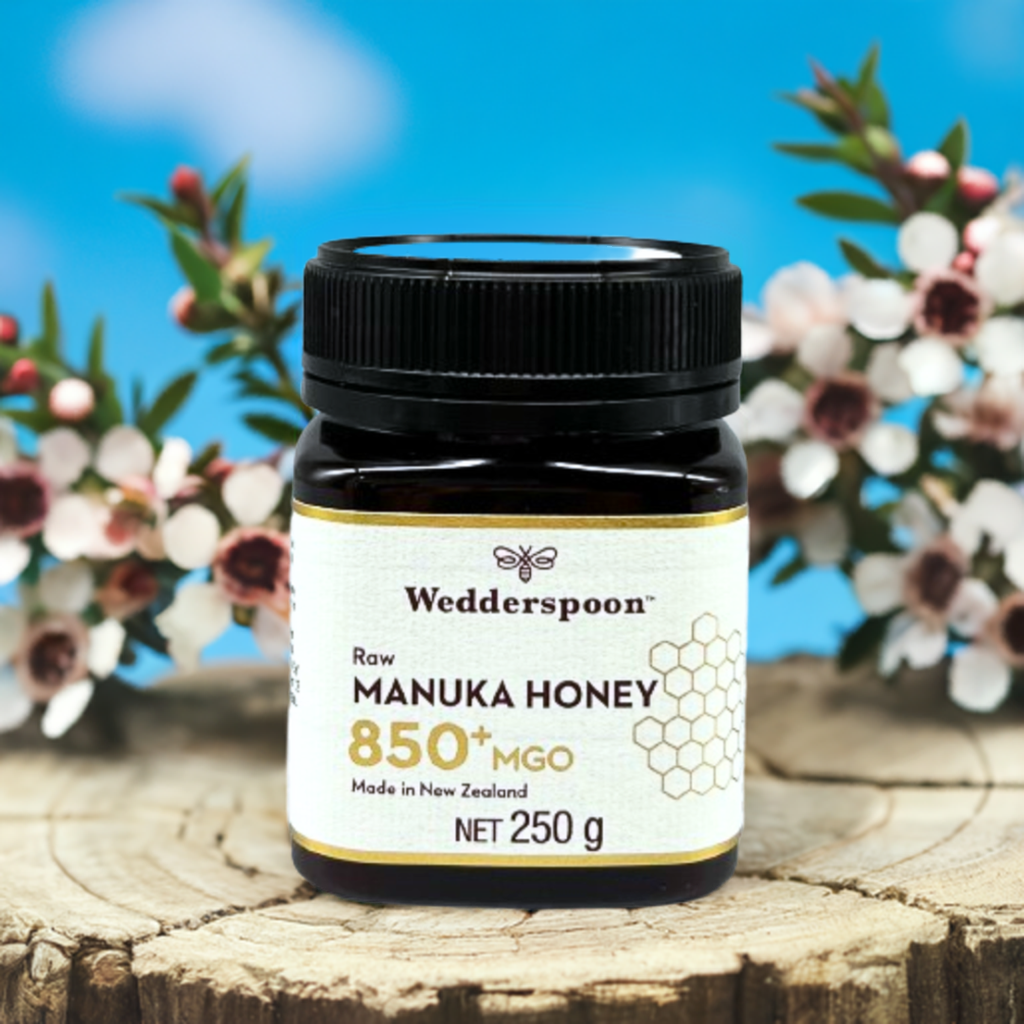Wedderspoon Raw Manuka Honey 850 MGO 250g . Dark glass jar on wooden stump with manuka flowers backdrop