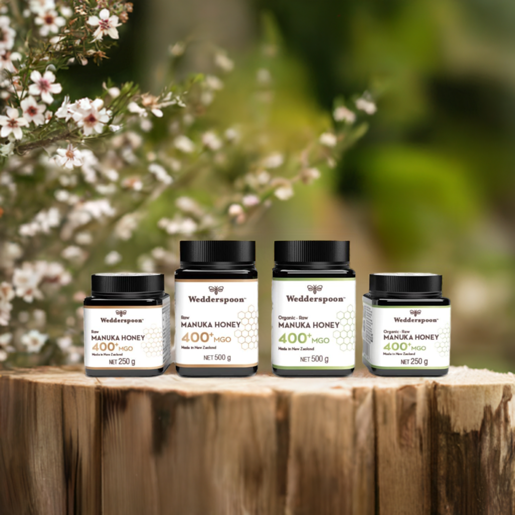 Wedderspoon Raw Manuka Honey 850 MGO 250g jar displayed with other Wedderspoon jars on wood stump and manuka blooms behind