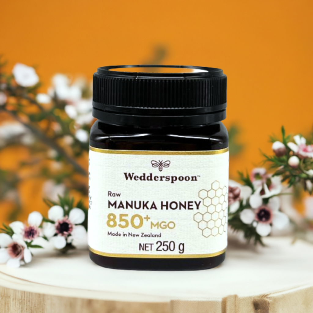 Wedderspoon Raw Manuka Honey 850 MGO 250g . dark glass jar on wooden board with label and manuka flowers in background