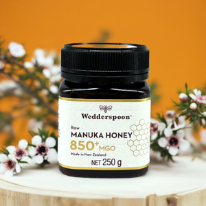 Wedderspoon Raw Manuka Honey 850 MGO 250g . dark glass jar on wooden board with label and manuka flowers in background