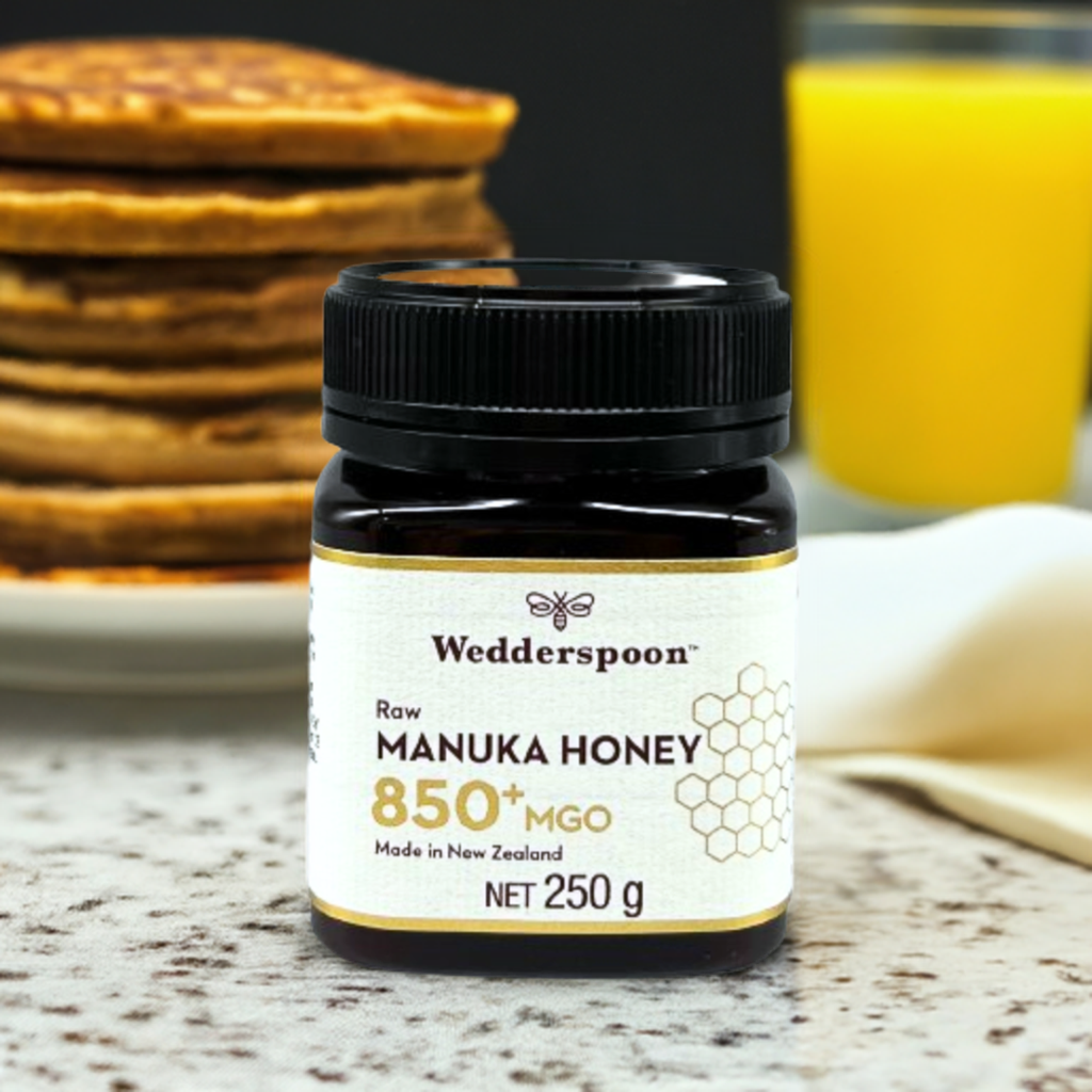 Wedderspoon Raw Manuka Honey 850 MGO 250g . Dark jar of Manuka honey on a kitchen table with pancakes and orange juice