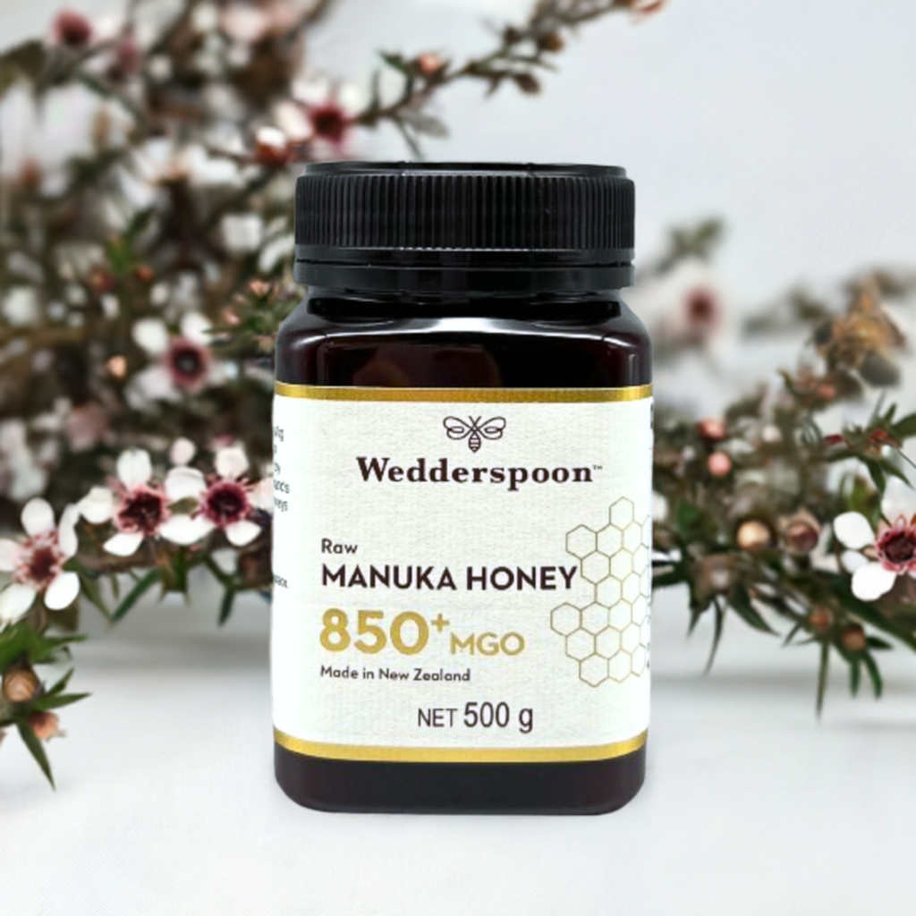 Wedderspoon Raw Manuka honey 850 MGO 500g jar on white surface with label visible and manuka flowers behind