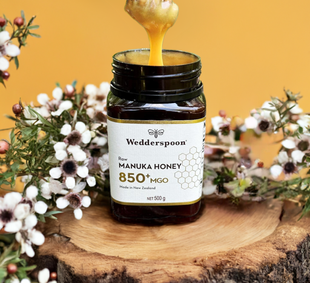 Wedderspoon Raw Manuka honey 850 MGO 500g. Jar open with honey drizzle on wood slice surrounded by manuka flowers