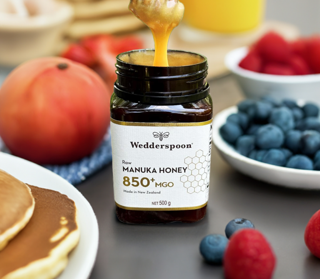 Jar of Wedderspoon Raw Manuka honey 850 MGO 500g being drizzled over pancakes in a breakfast setting with berries