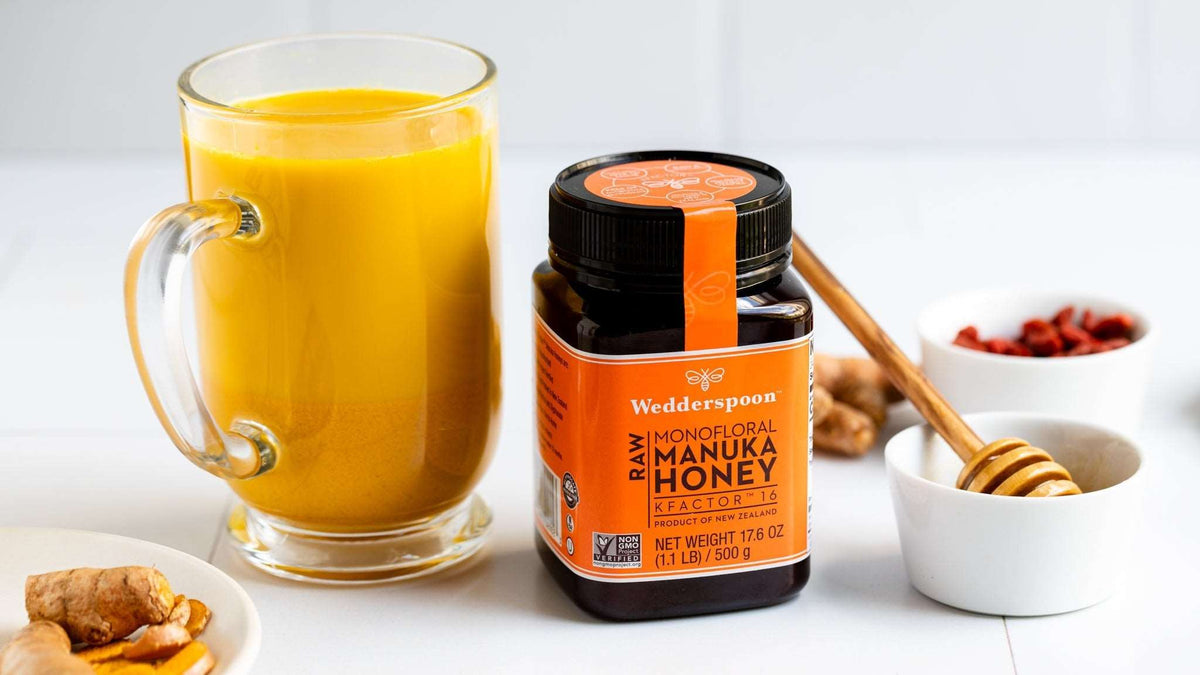 Wedderspoon Raw Manuka Honey KFactor 16 500g Triple Pack jar beside a glass of turmeric tea and honey dipper