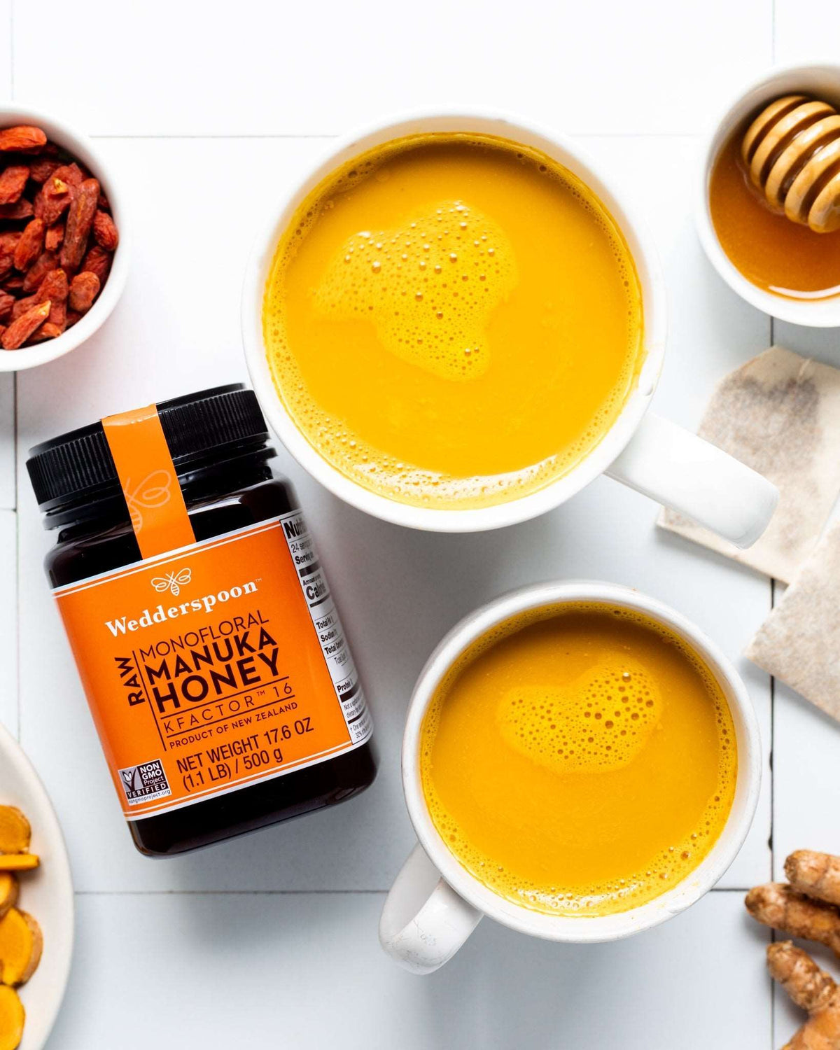 Wedderspoon Raw Manuka Honey KFactor 16 500g Triple Pack jar beside two cups of tea with honey dipper and natural ingredients